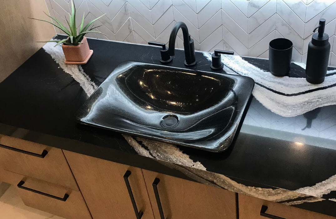 Modern bathroom countertop featuring a black square vessel sink with matte black faucet and accessories, accented by a natural stone pattern and a small potted plant.