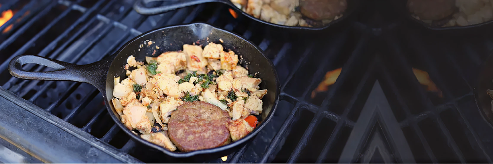 Cast iron skillet on a grill with cooked scrambled eggs mixed with herbs and diced potatoes, accompanied by a sausage patty.