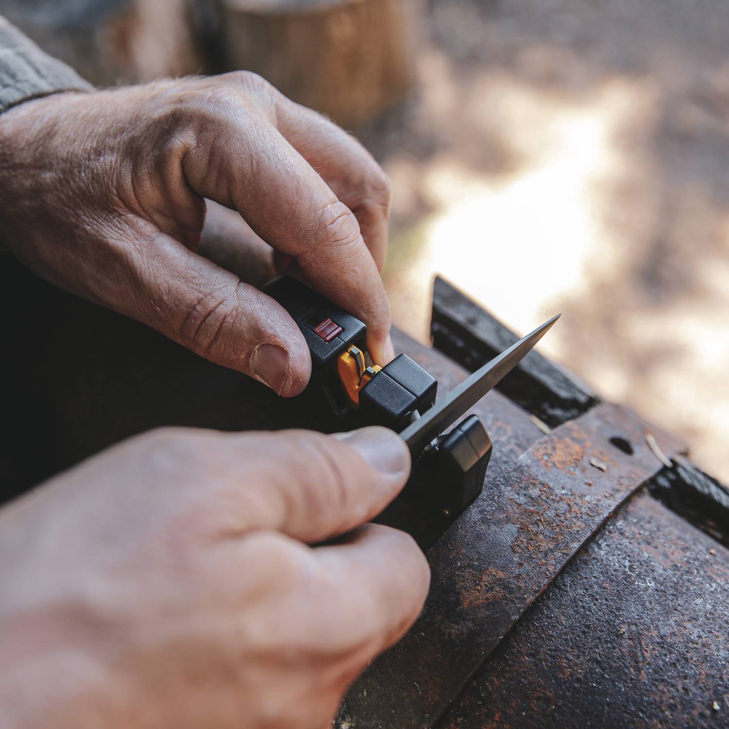Hands sharpening a knife using a black and yellow Work Sharp Pivot Plus tool on a rustic metal surface