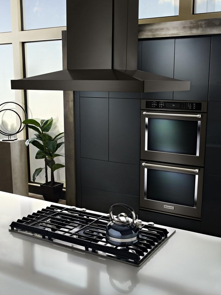 Matte black island-mount canopy hood above a modern gas cooktop with stainless steel kettle, set against dark cabinetry in a bright kitchen, featured in Island Range Hoods and Kitchen & Kitchenware collections.