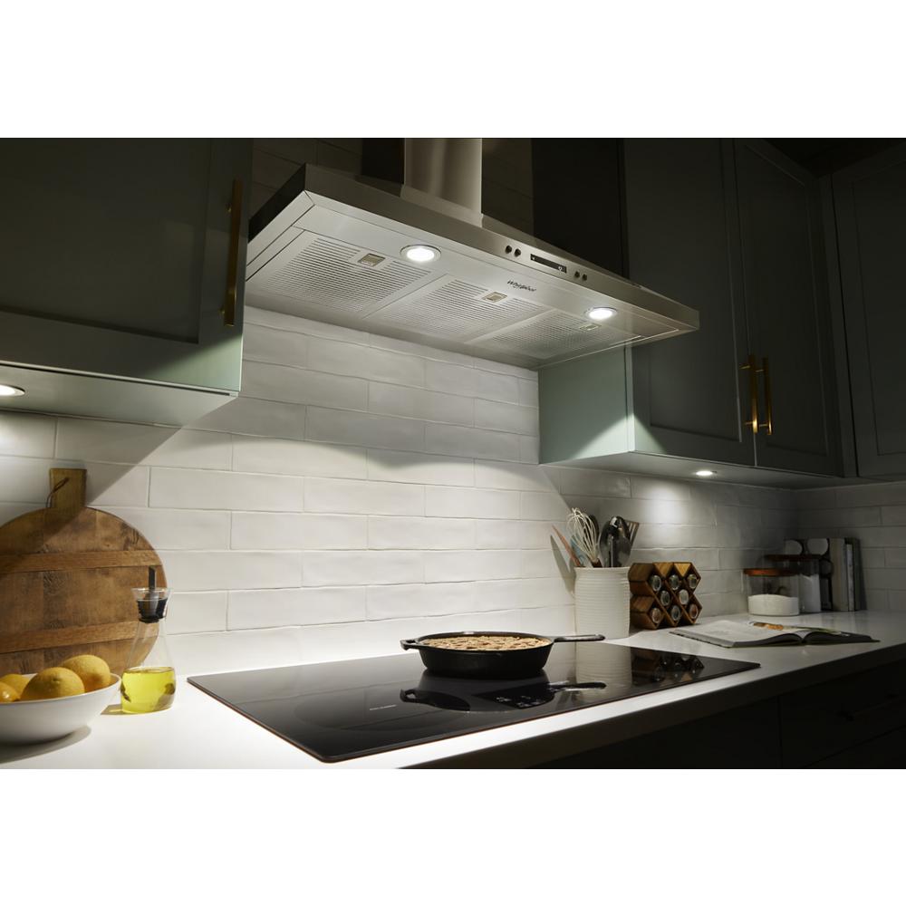 Illuminated stainless steel wall mount chimney range hood over induction cooktop in modern kitchen setting