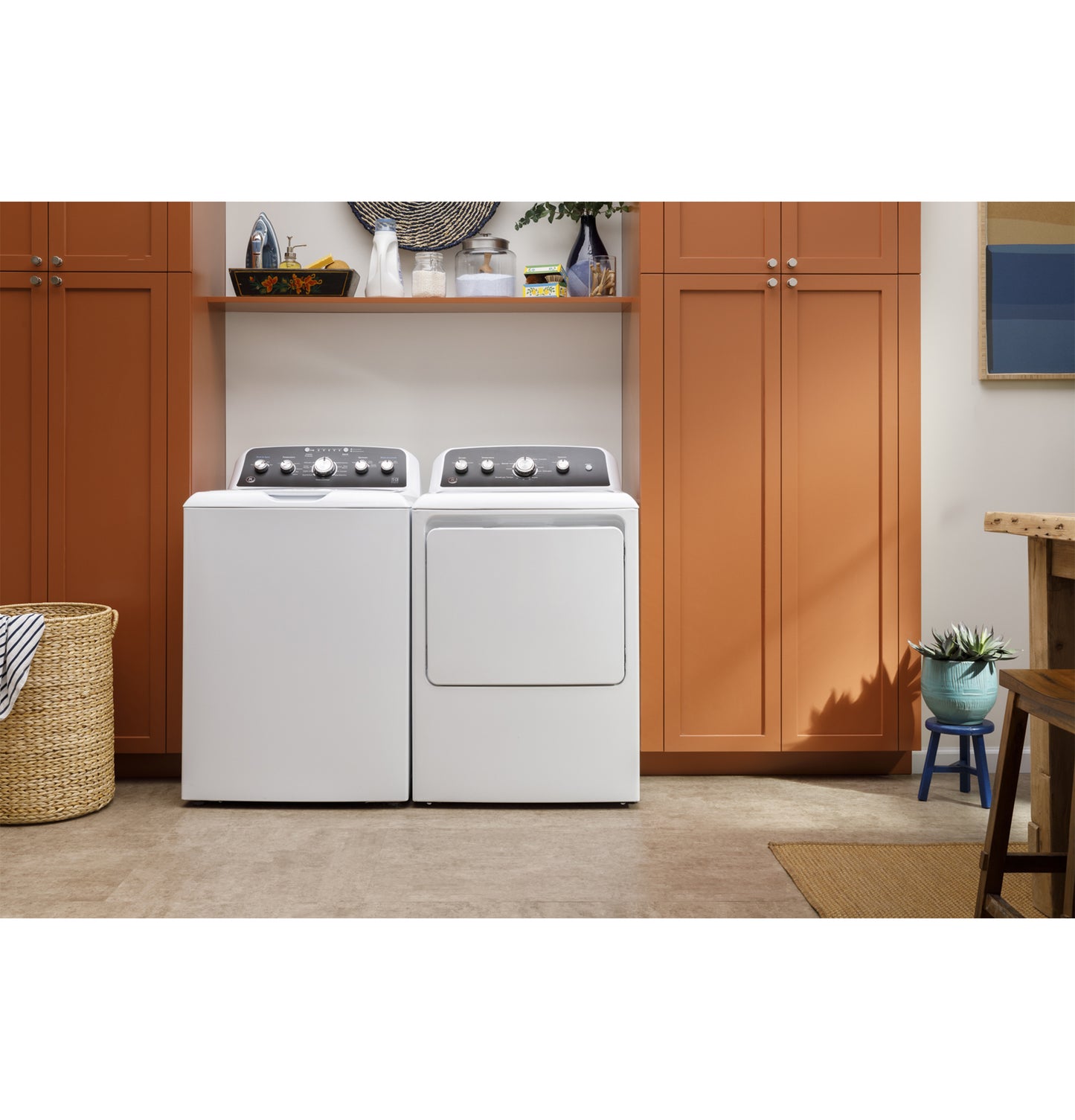 White top load washer and dryer set with dark control panels in a laundry room with orange cabinets and woven basket nearby