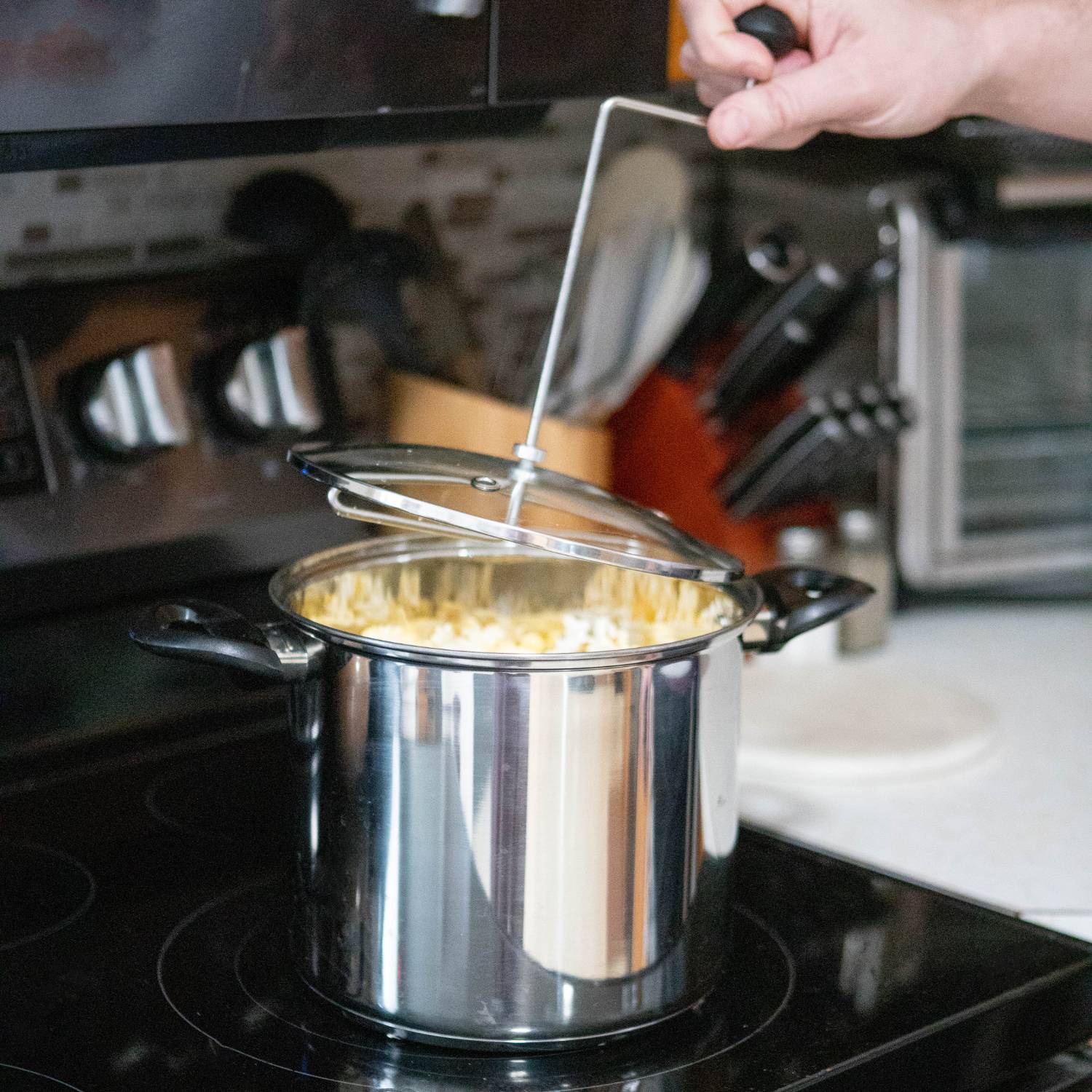 Hand turning crank on stainless steel popcorn popper with glass lid on stovetop in kitchen setting