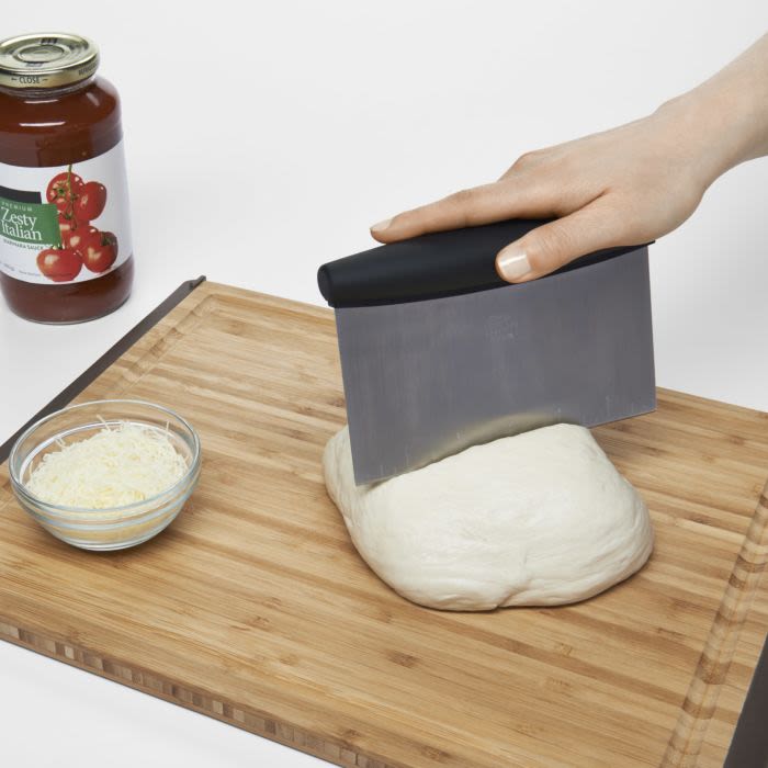 Hand gripping black-handled stainless steel scraper cutting dough on wooden board with cheese bowl and jar nearby in kitchenware setting