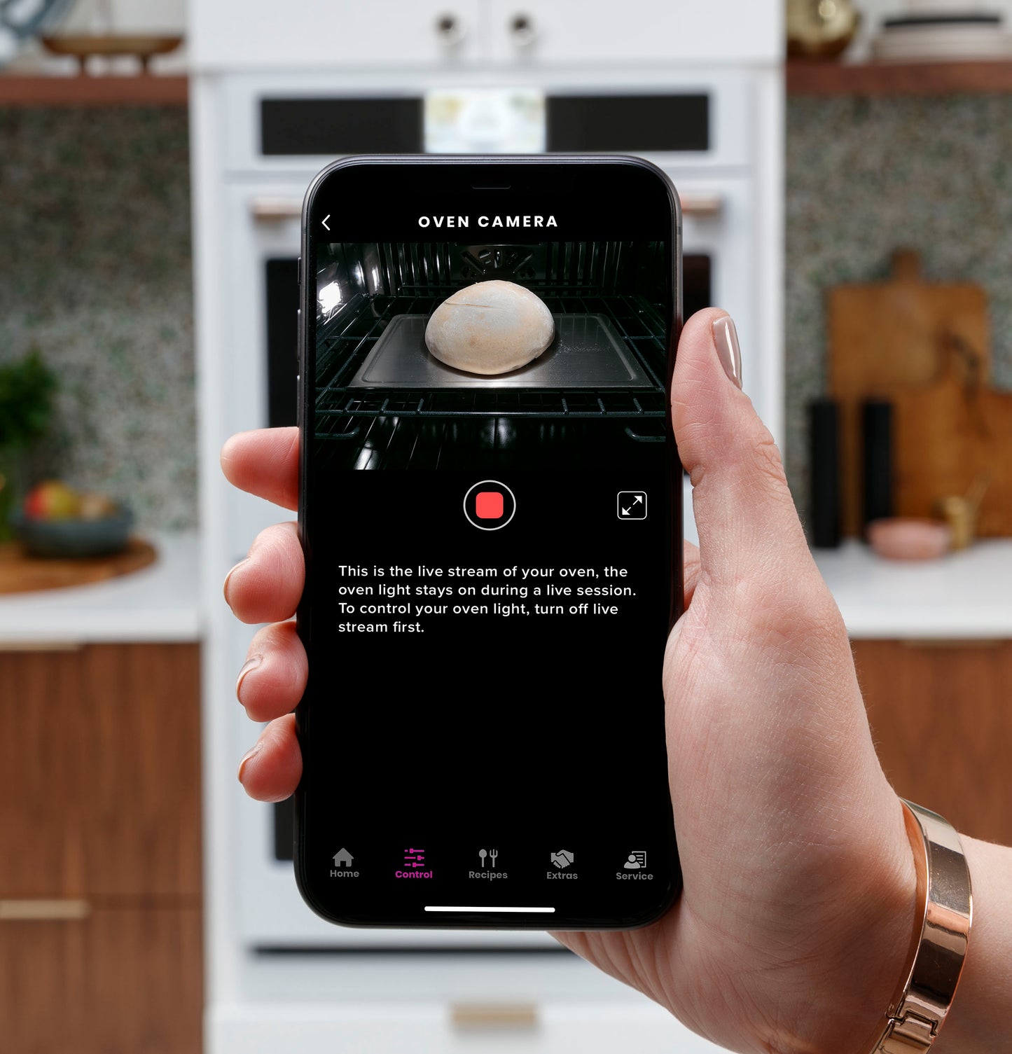 Hand holding smartphone showing live oven camera feed of bread baking inside built-in wall oven in modern kitchen