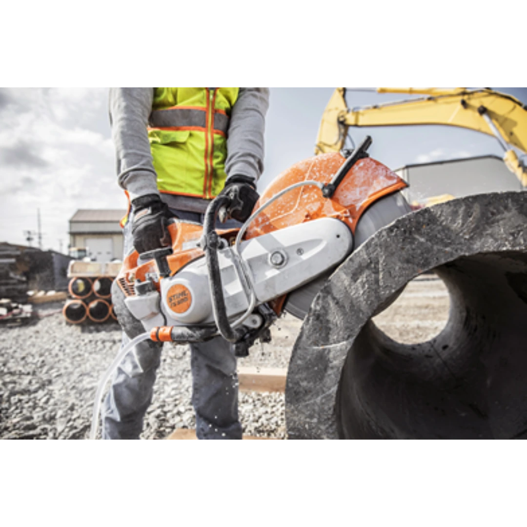 Worker operating an orange STIHL power cutter with water hose to cut large concrete pipe at construction site