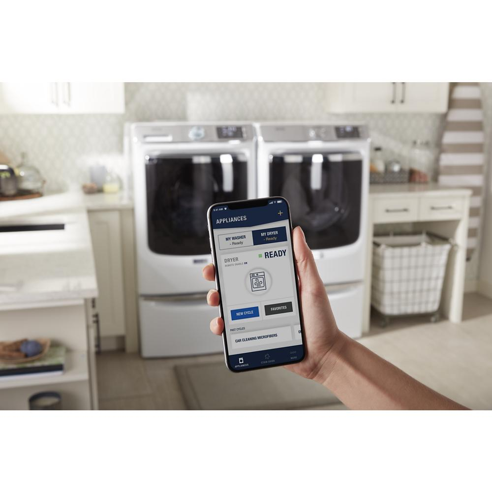 Hand holding smartphone displaying smart dryer app with front load dryer and washer in bright laundry room setting