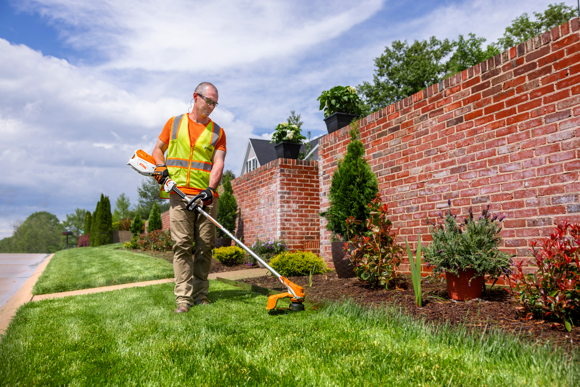 Man wearing safety vest using STIHL battery-powered trimmer along garden bed for outdoor lawn and home improvement tasks