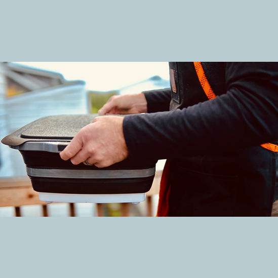 Person holding collapsible black BBQ prep tub with lid, ideal for grilling and outdoor living tasks under $100