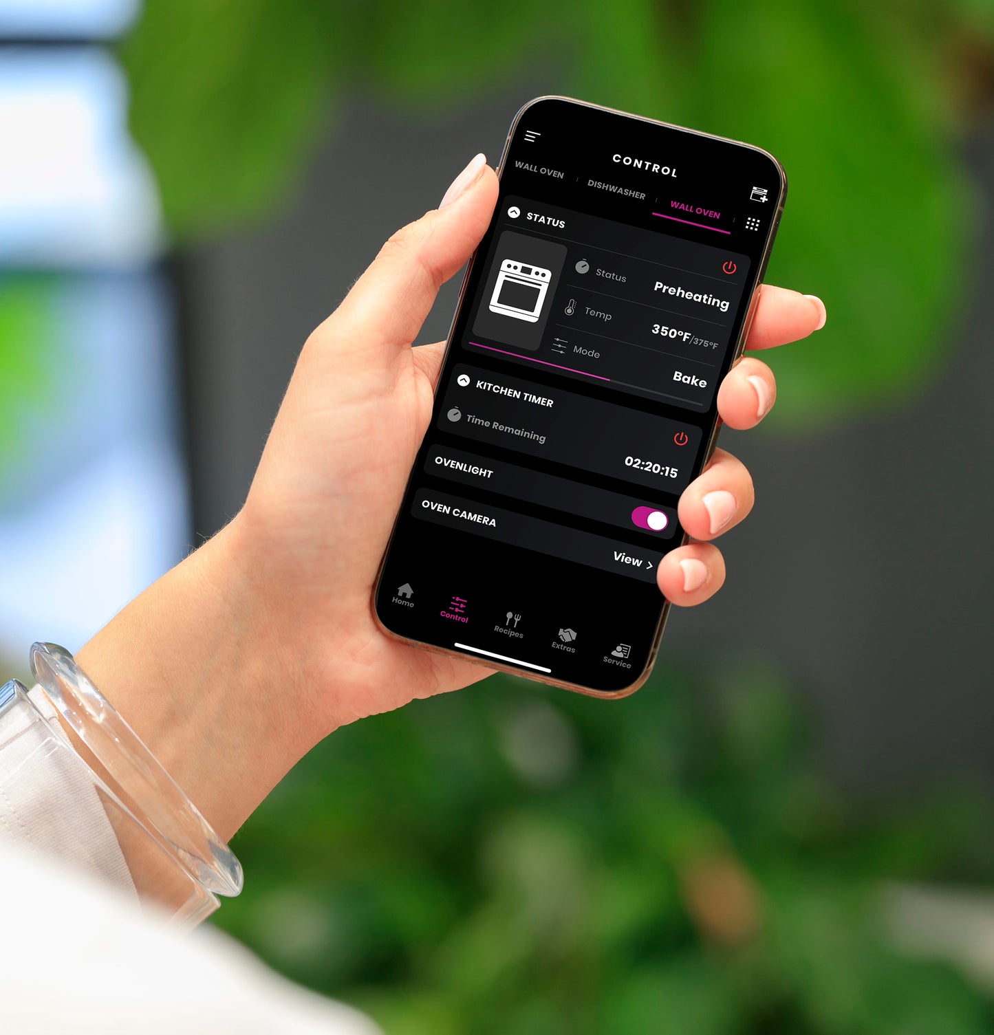 Hand holding smartphone displaying smart control app preheating built-in wall oven at 350 degrees in modern kitchen setting