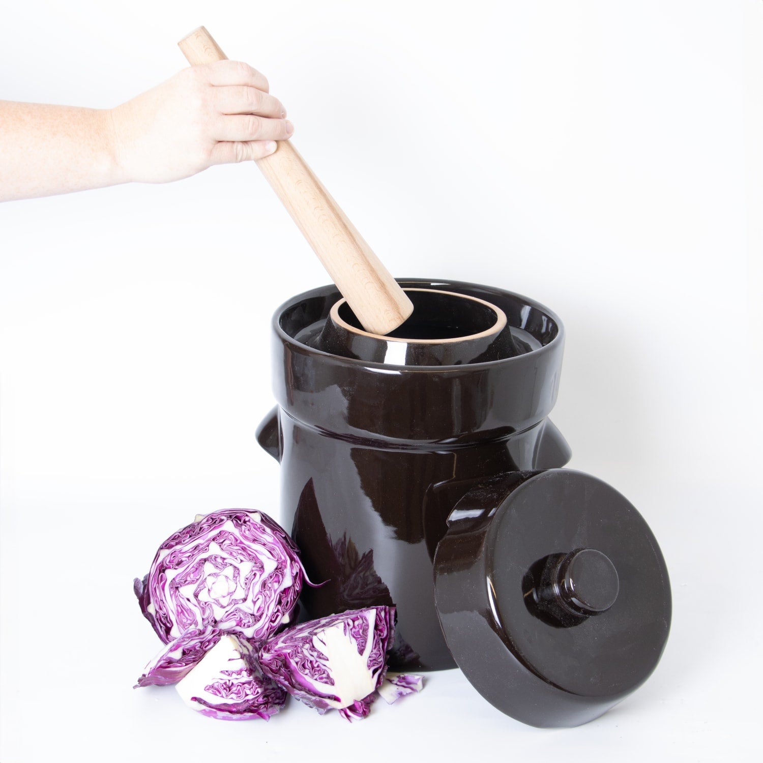 Hand pressing wooden stomper inside glossy black crock with lid nearby and cut purple cabbage on white surface