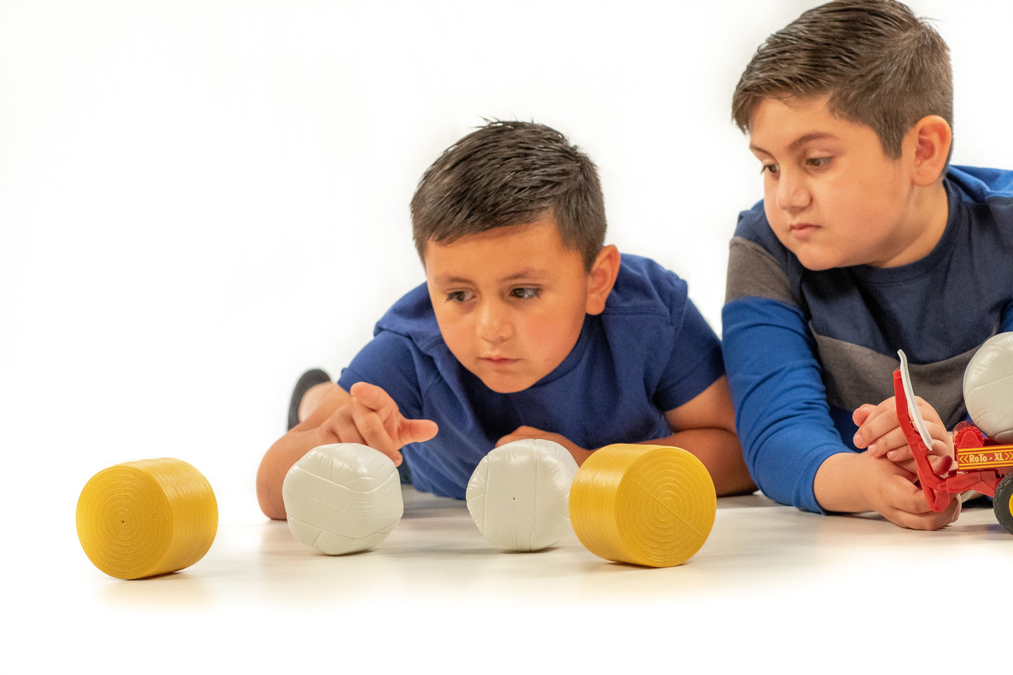 Two boys playing with yellow and white plastic round bale toys on a white surface for farm play