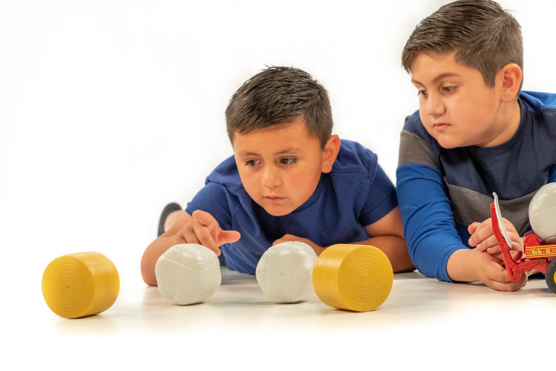 Two boys playing with yellow and white plastic round bale toys on a white surface for farm play