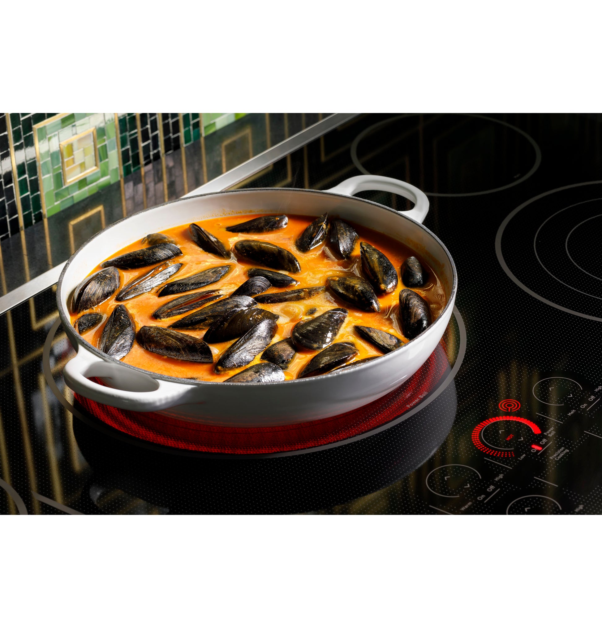 White ceramic pan simmering mussels in broth on a black glass touch-control cooktop with green tiled backsplash