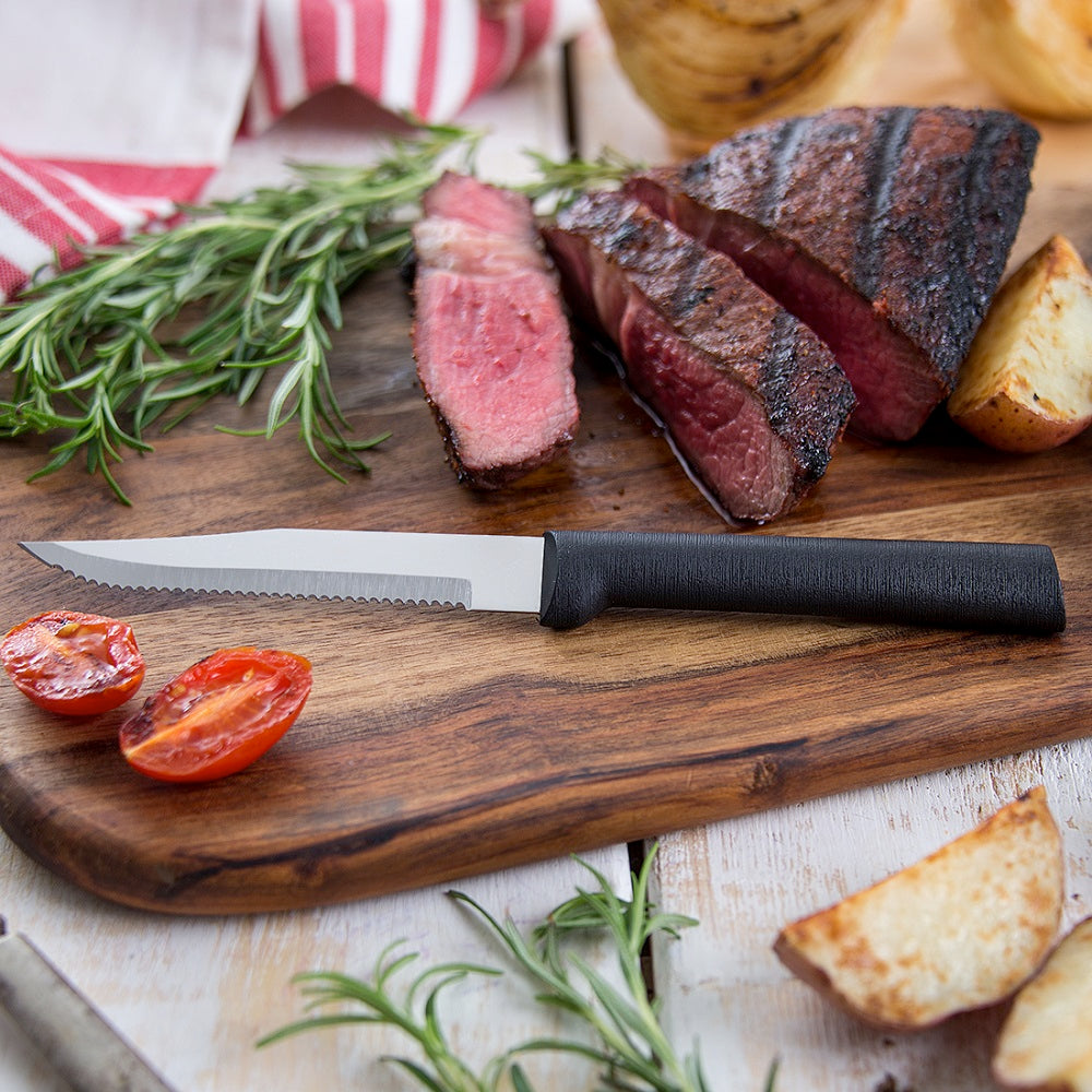 Steak knife with serrated blade and black handle on wooden board with grilled meat and roasted potatoes in kitchenware collection