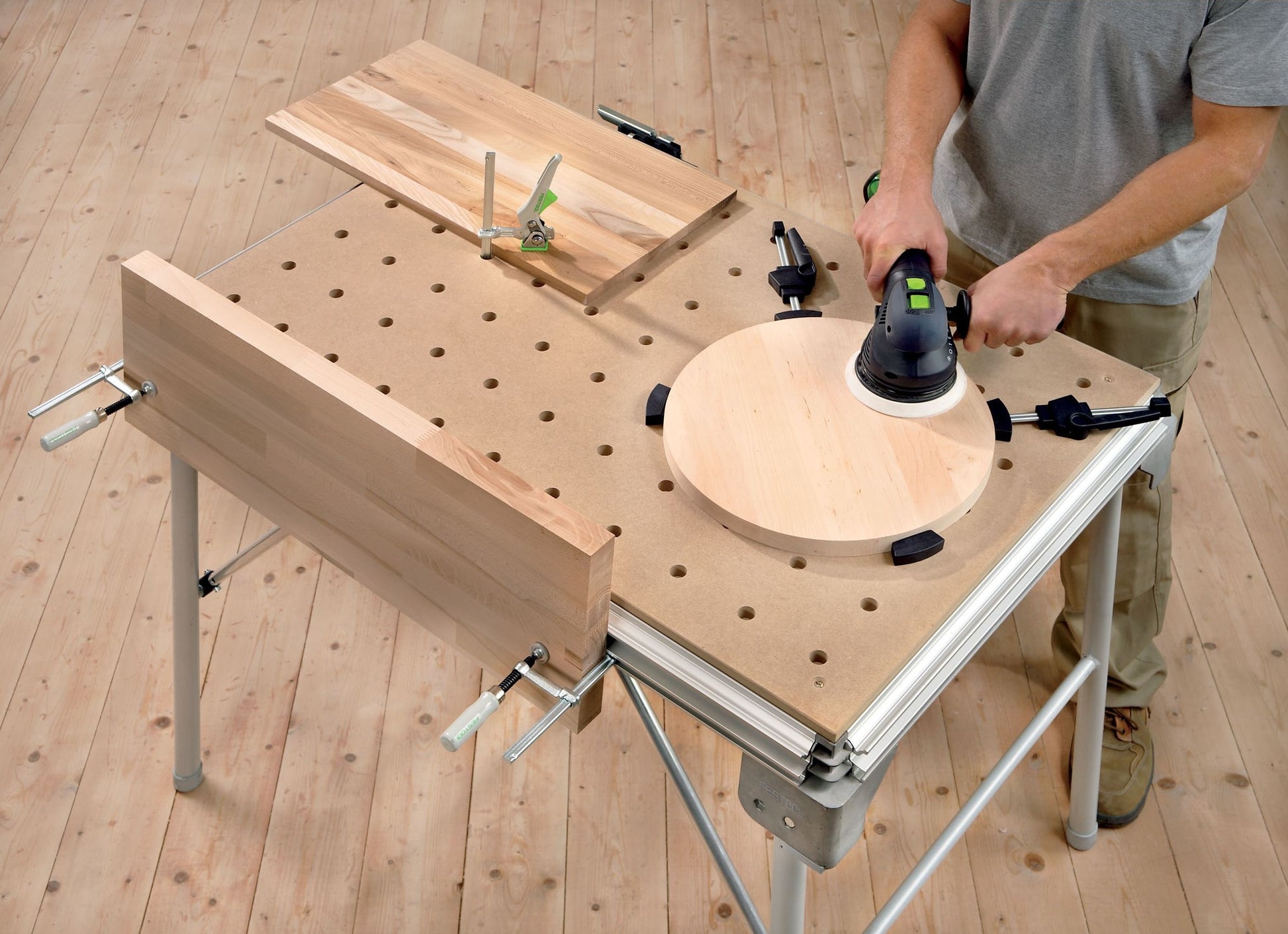 Multifunction woodworking table with clamped wooden pieces and a person sanding a round wood surface using a power tool.