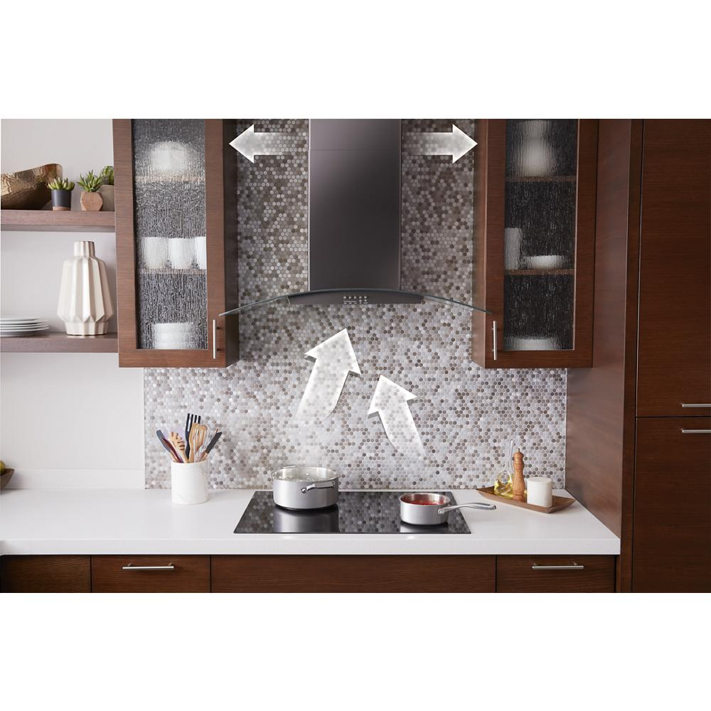Modern kitchen with a curved glass wall mount range hood ventilating over a cooktop amidst wood cabinetry and mosaic tile backsplash