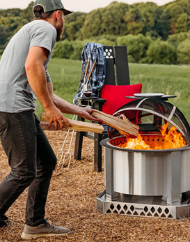 Man placing wood logs into a stainless steel smokeless fire pit in an outdoor backyard setting for home improvement use