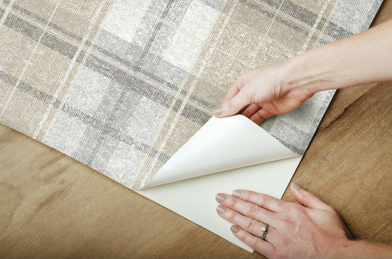 Hands applying gray and beige tweed plaid peel and stick wallpaper on wooden surface for home improvement
