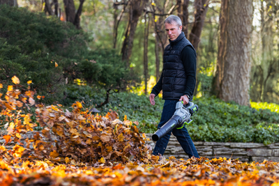 Man using EGO Power+ cordless leaf blower with battery to clear fallen leaves in a backyard garden setting
