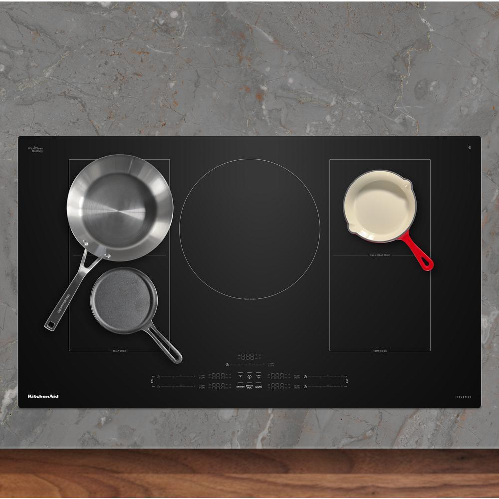Black induction cooktop with five cooking zones holding stainless steel, cast iron, and ceramic skillets in kitchenware setting