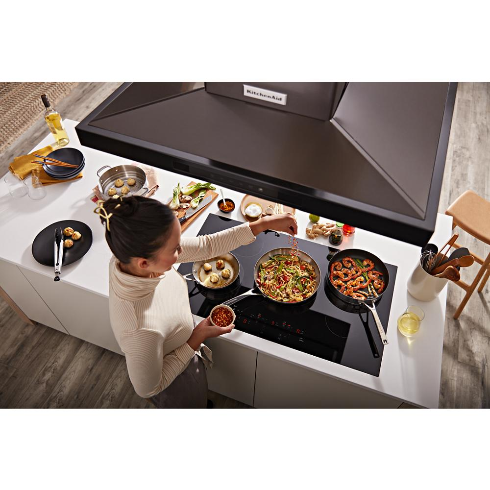 Woman cooking multiple dishes on black cooktop under a dark stainless steel canopy hood from KitchenAid range hoods collection