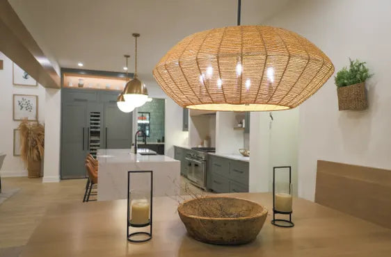 Dining area featuring a large woven pendant light above a wooden table set with decorative candles and a rustic bowl, adjacent to a kitchen with modern cabinetry.