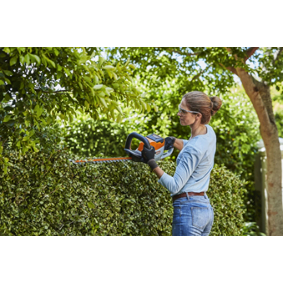 Woman wearing safety glasses trimming green hedge with lightweight battery-powered orange and white hedge trimmer outdoors