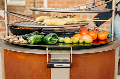 Round hot-rolled carbon steel griddle with dual handles cooking vegetables and corn over outdoor fire pit setting