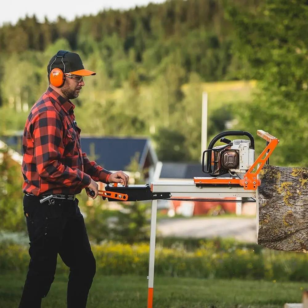 Man in ear protection and plaid shirt operating orange and black crank feed attachment for gas saw in outdoor setting