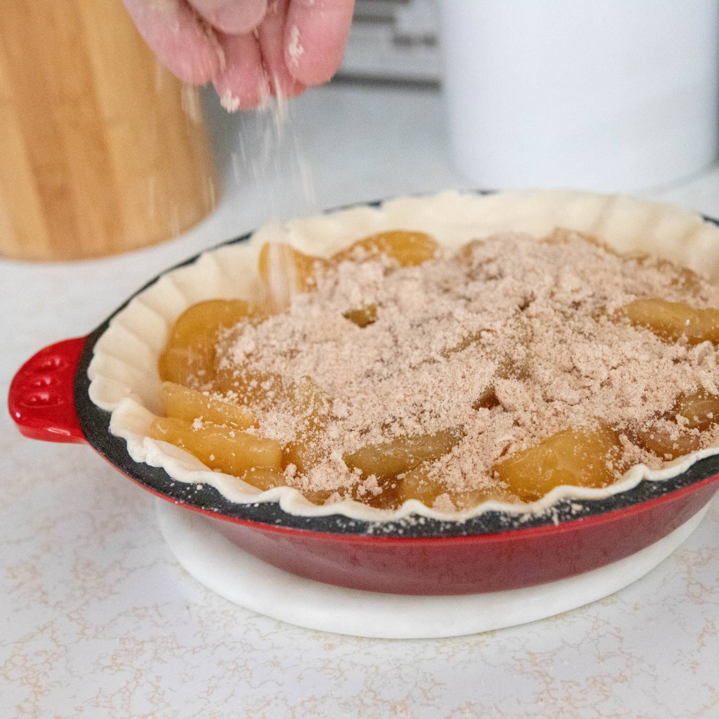 Hand sprinkling cinnamon sugar topping over uncooked apple pie filling in red cast iron pan with crimped edges