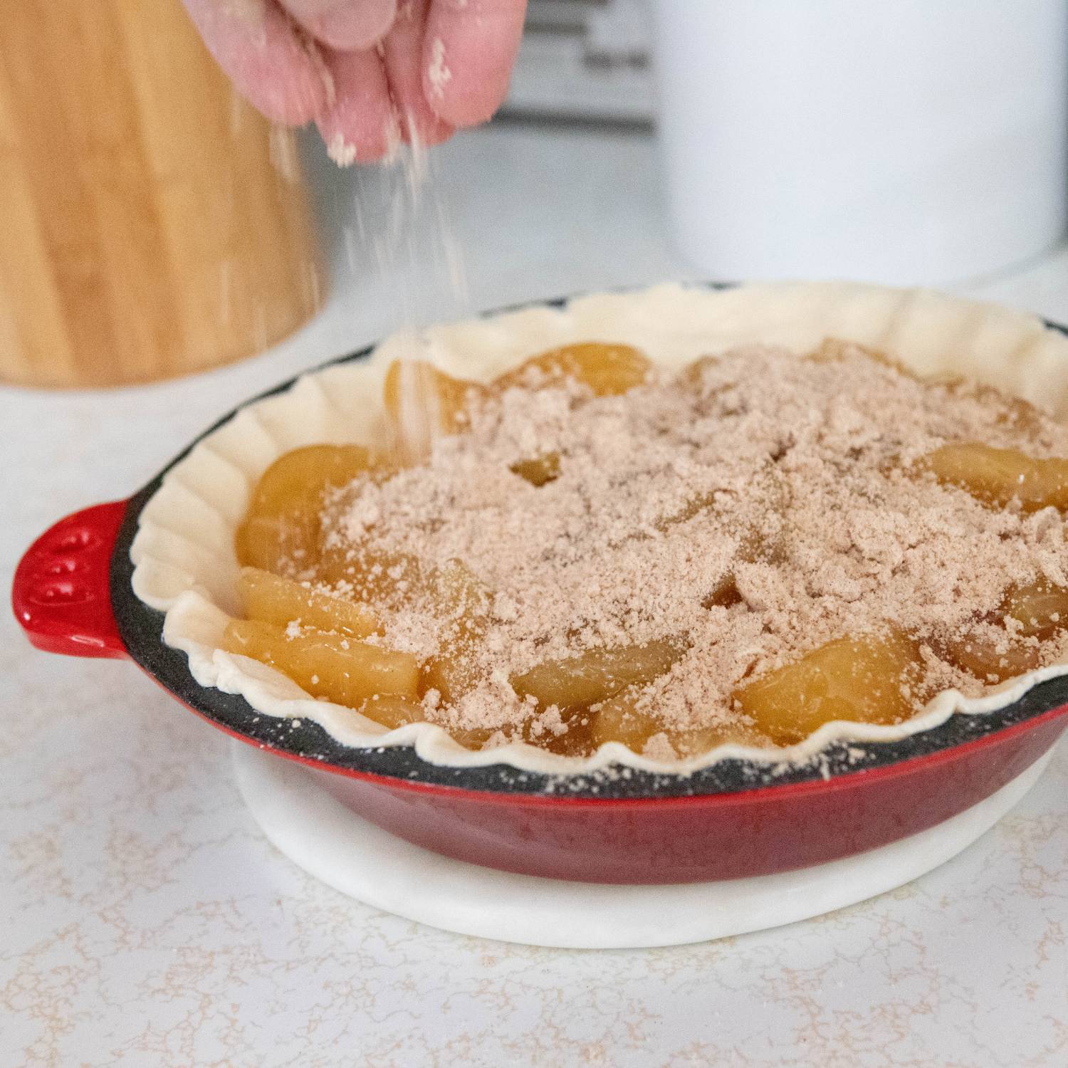 Hand sprinkling cinnamon sugar topping over uncooked apple pie filling in red cast iron pan with crimped edges