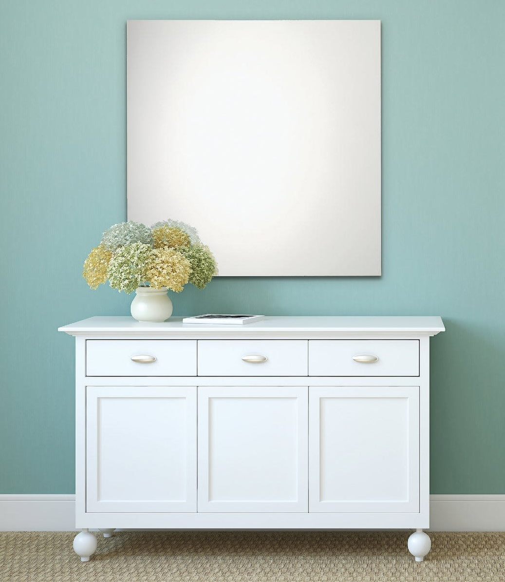 Square frameless wall mirror with polished edges above white cabinet adorned with floral arrangement and small book