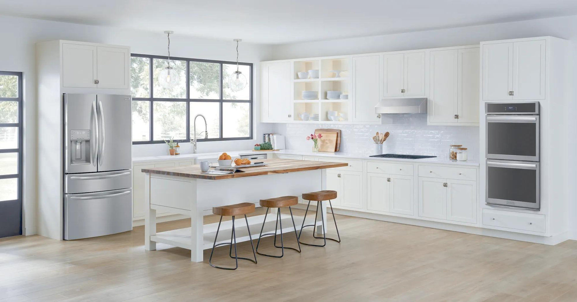 Modern white kitchen featuring stainless steel double electric wall oven, French door refrigerator, and island seating in bright space