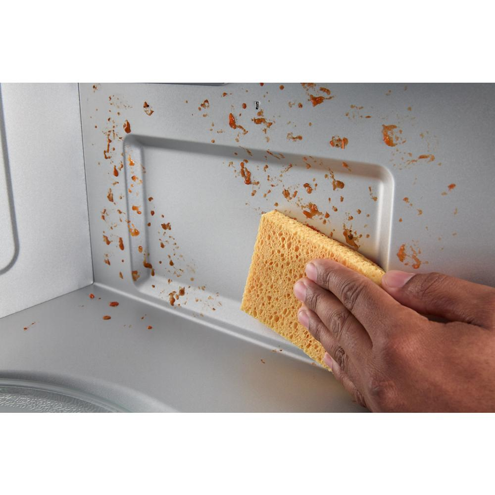 Hand using yellow sponge to clean food splatters inside microwave cavity for kitchen appliance maintenance
