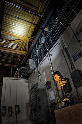 Yellow rechargeable LED spotlight mounted on a stand illuminating an industrial workspace with exposed pipes and panels