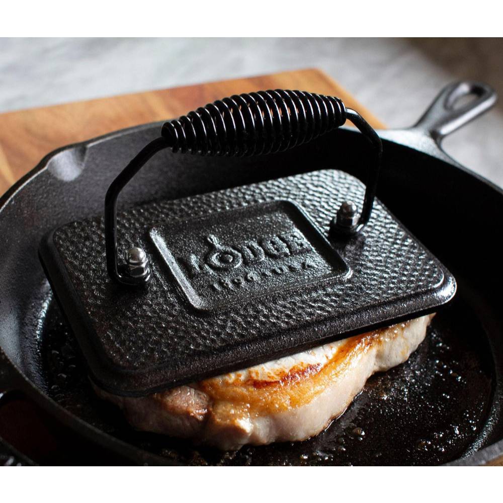 Square cast iron grill press with coil handle resting on a searing piece of meat in a cast iron skillet under $50 kitchenware collection