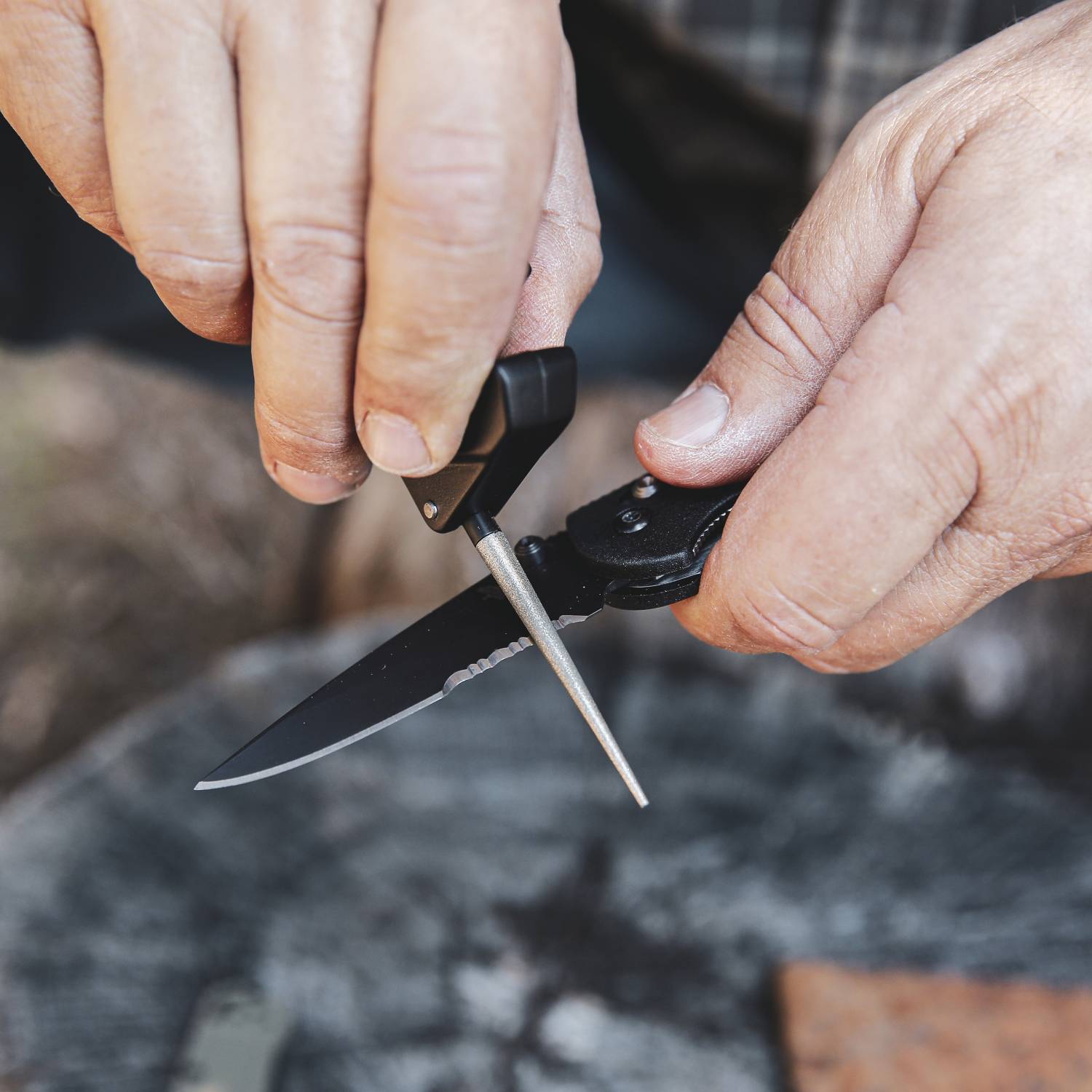 Person using a handheld sharpener with a metal rod to maintain edge on a folding serrated knife outdoors