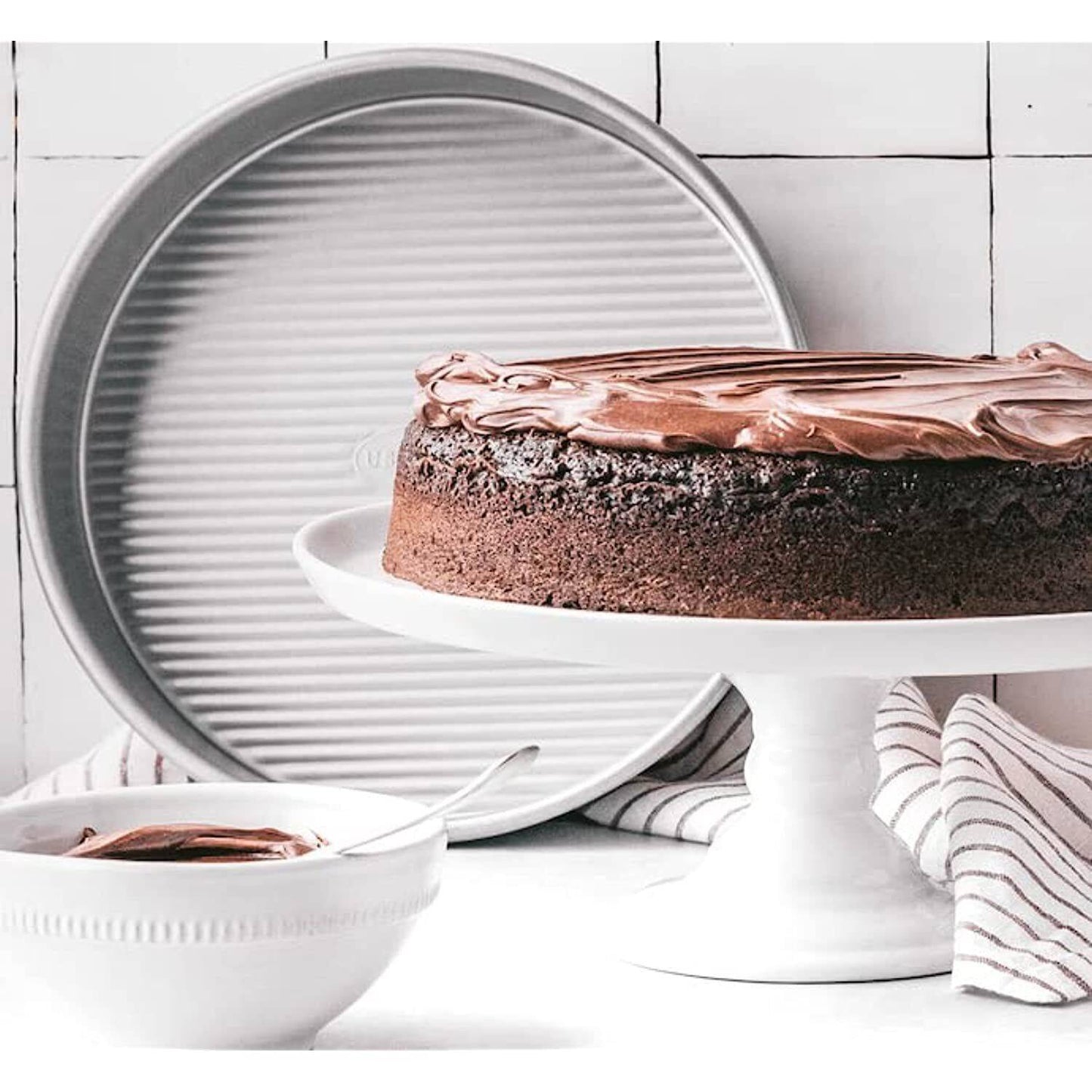 9-inch round cake pan with ridged interior behind chocolate frosted cake on white pedestal with striped cloth and bowl