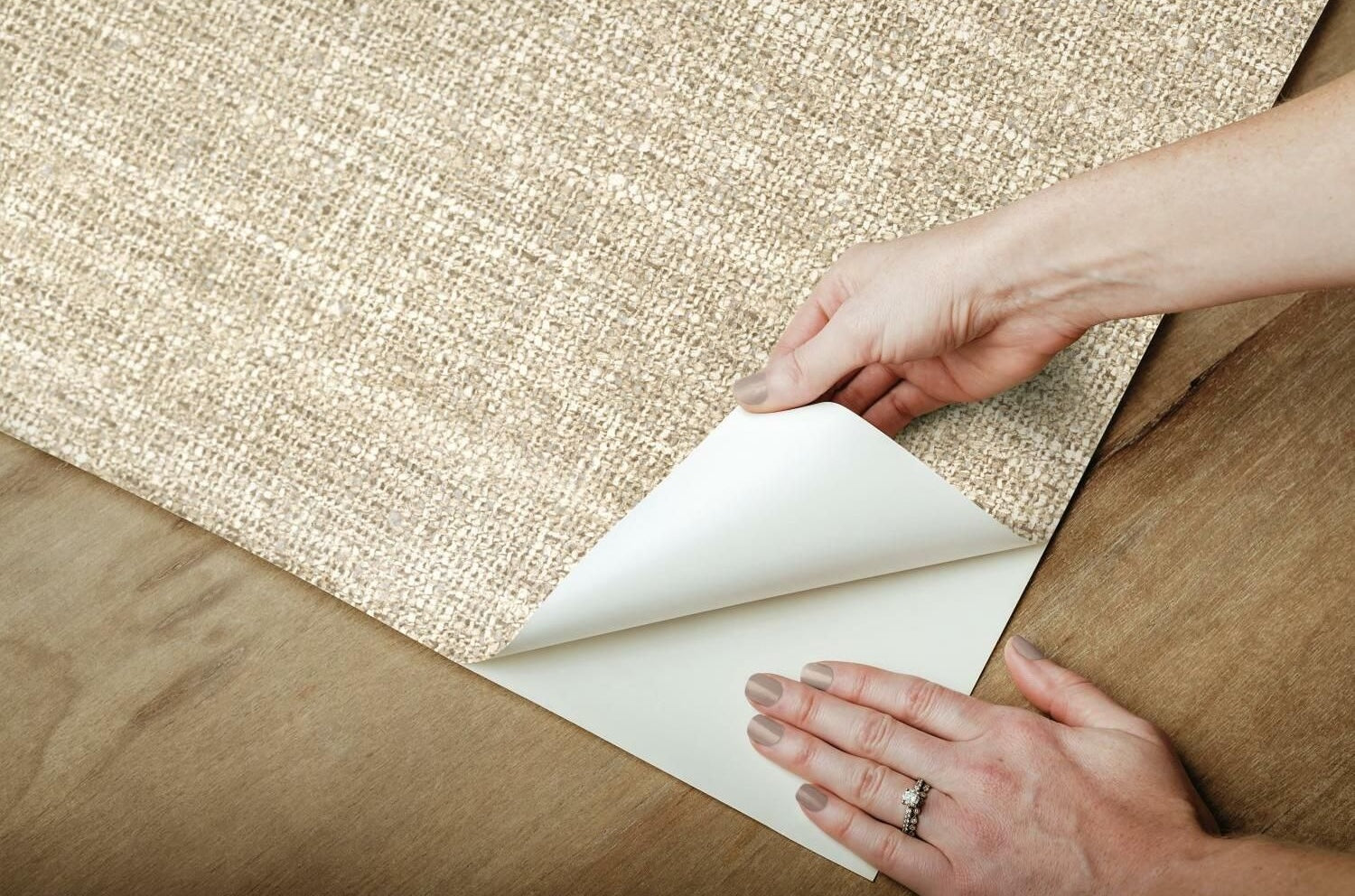 Hands peeling back brown tweed textured wallpaper with adhesive backing on wood surface for home improvement