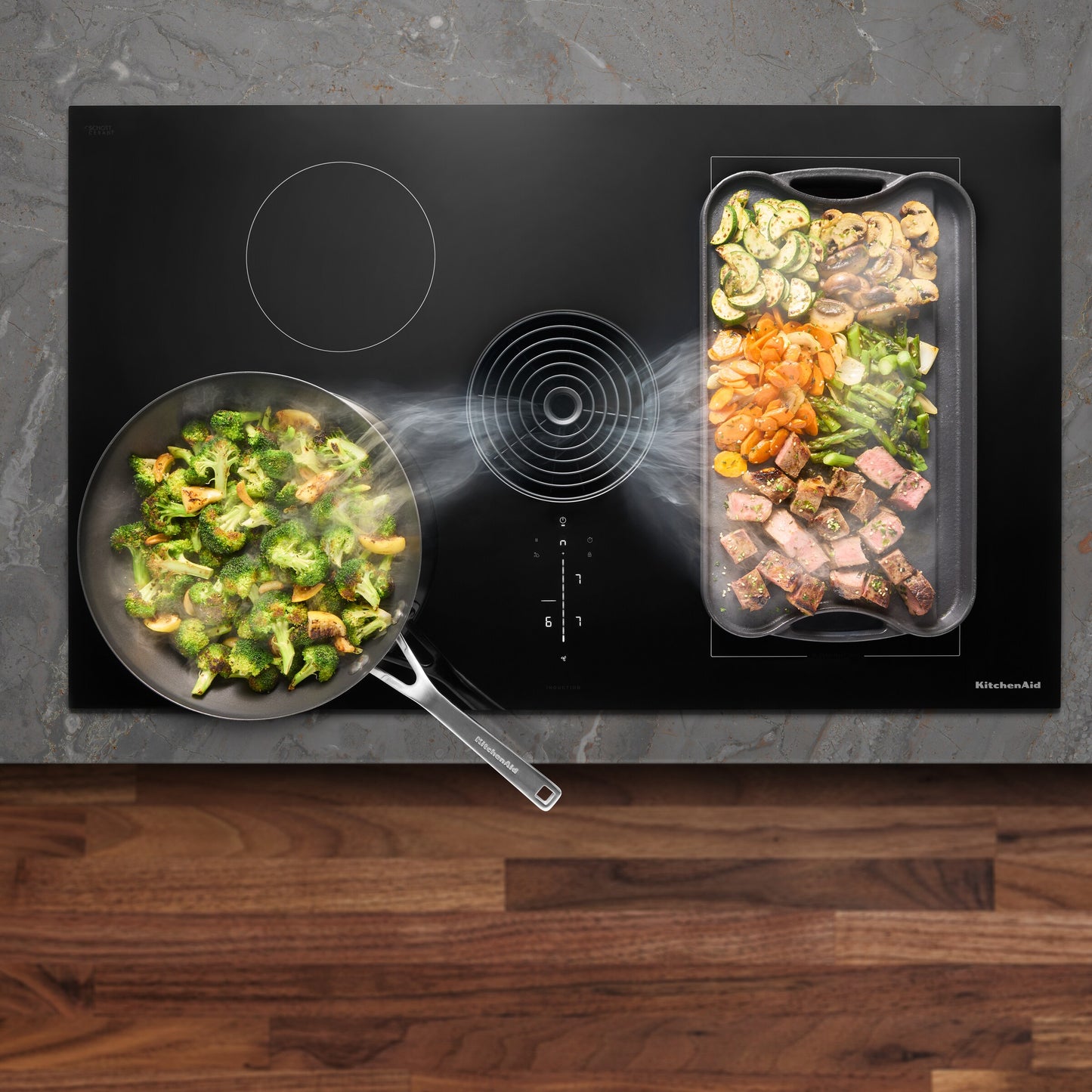 Overhead view of black induction cooktop with downdraft vent steaming while vegetables and meat cook in pans