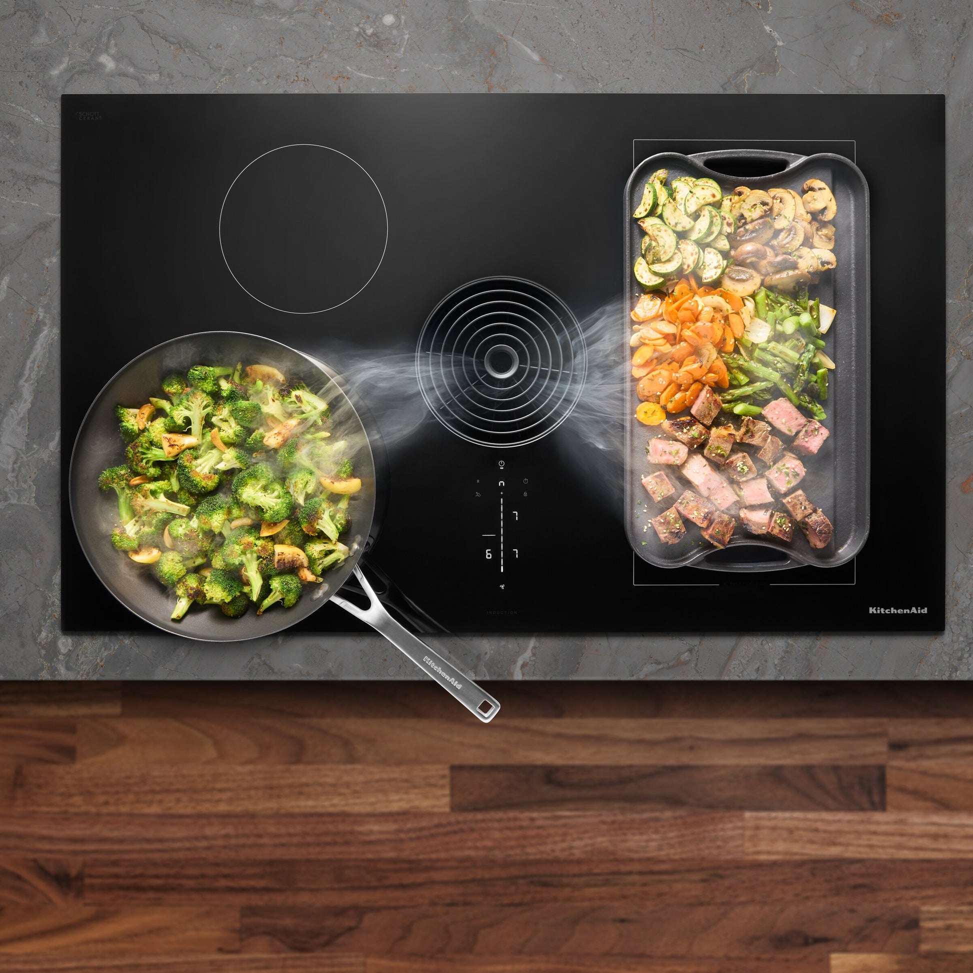 Overhead view of black induction cooktop with downdraft vent steaming while vegetables and meat cook in pans