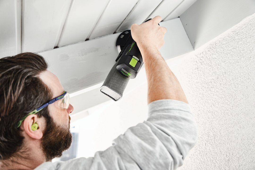 Man wearing safety glasses using a cordless Festool sander on overhead wooden beams for home improvement tasks