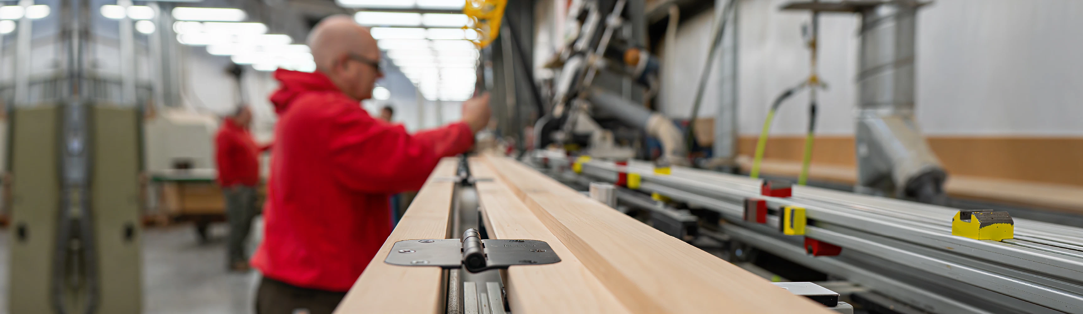 Worker in a red sweatshirt operating machinery to process long wooden boards inside a well-lit woodworking facility focused on precision and safety.