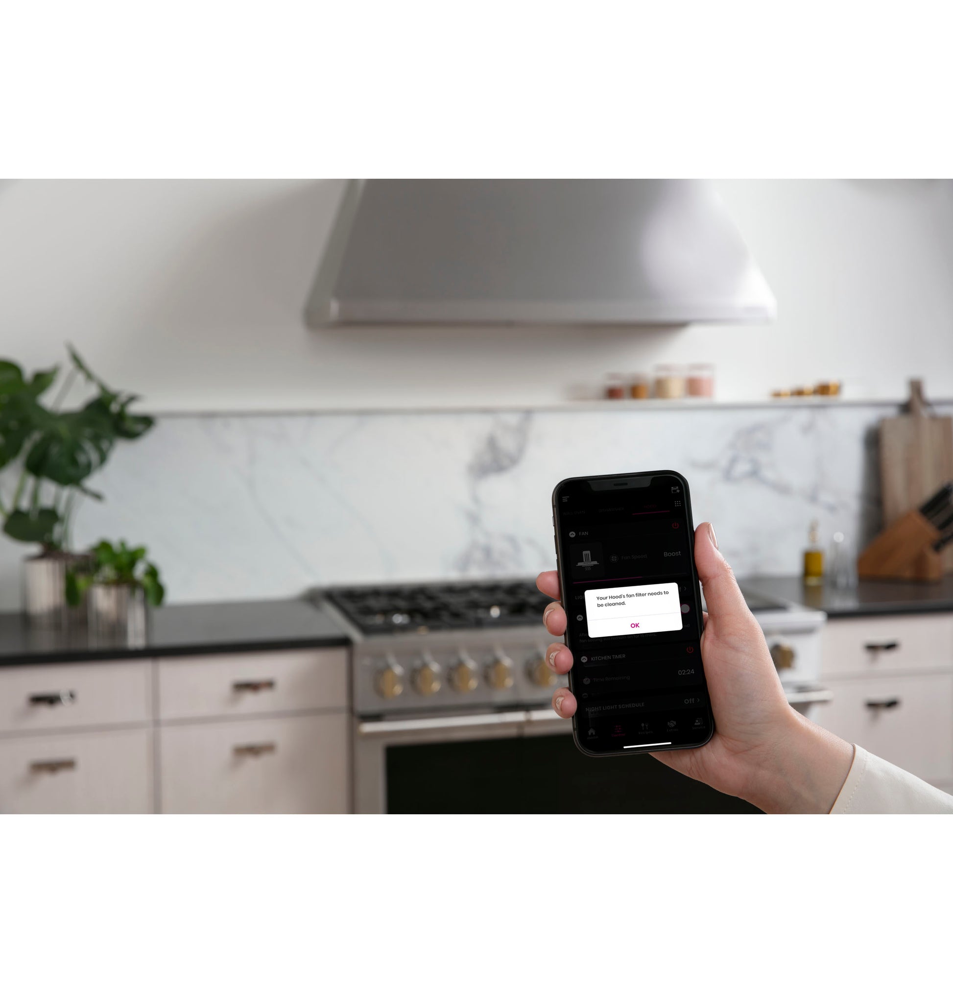 Hand holding smartphone controlling modern stainless steel range hood above gas stove in kitchen appliances setting