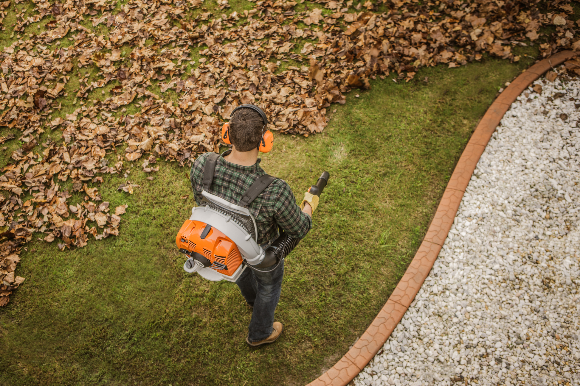 Person using orange backpack blower with ear protection to clear leaves on lawn near stone border for home improvement tasks