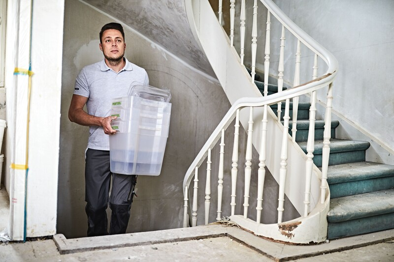 Man carrying clear plastic storage container up worn indoor staircase in home improvement setting