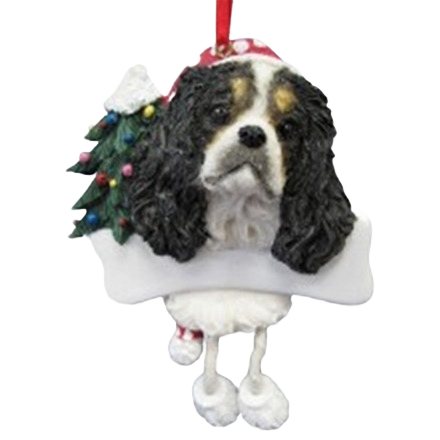 Tricolor King Charles dog ornament with dangling legs, wearing Santa hat next to decorated Christmas tree