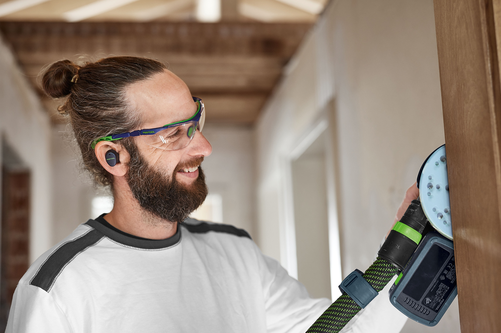 Man using cordless random orbit sander with safety glasses and earbuds in woodworking power tools and home improvement setting