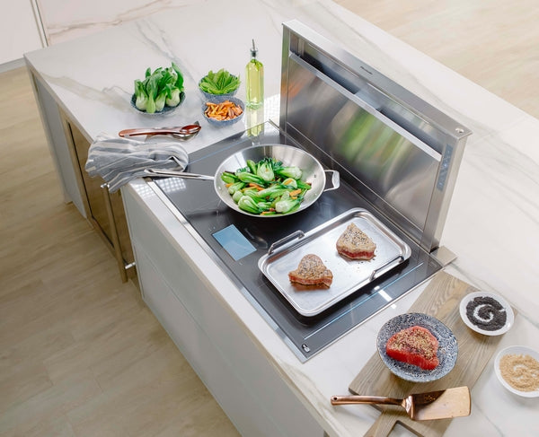 Stainless steel downdraft ventilation system integrated into kitchen island with cooking utensils and fresh vegetables nearby